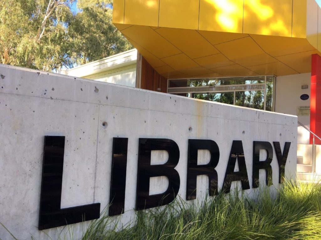 The word library in black capital letters against a pale grey wall with a yellow awning in the background