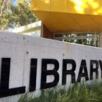 The word library in black capital letters against a pale grey wall with a yellow awning in the background