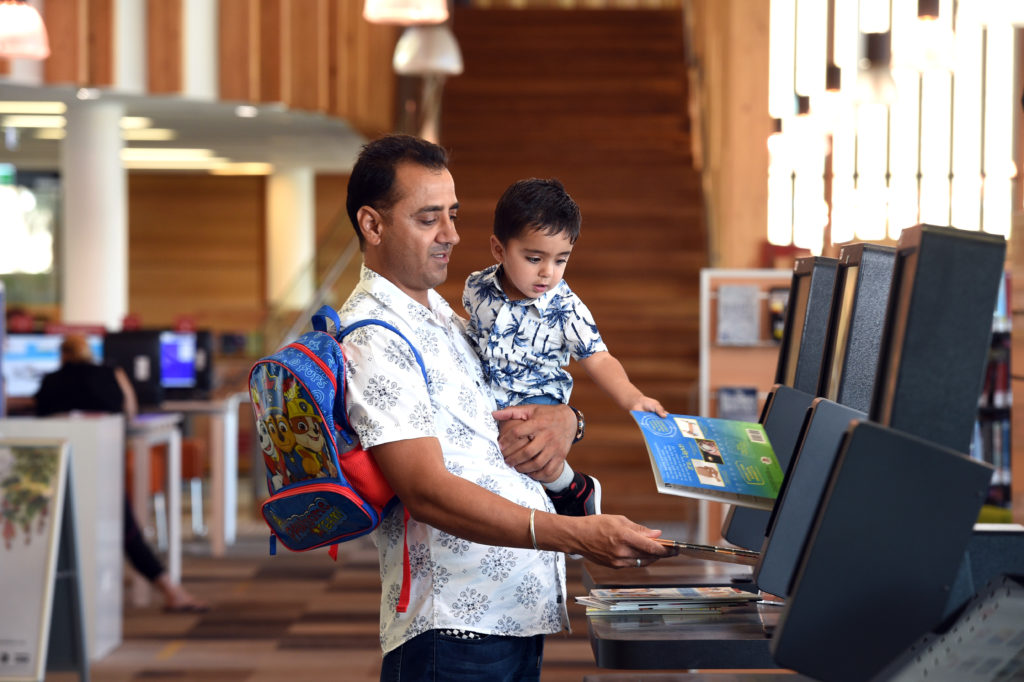 A man in a white short with a brightly coloured child's backpack over one shoulder holds his son, who is holding a book against the library check out kiosk.