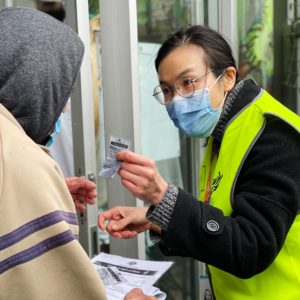 Woman wearing glasses, blue face mask and hi-vis vest hands vaccination information to a person wearing a hoodie