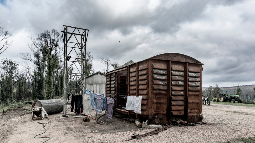 Photograph of burned out railway carriage beneath a grey cloudy sky with clothes drying on line in forefront