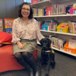 A smiling woman sits on a couch beside a black labrador wearing a seeing eye dog harness. In the background are shelves stacked with picture books as in a children's library.