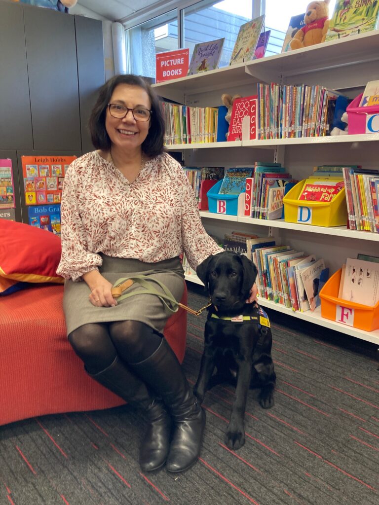 A smiling woman sits on a couch beside a black labrador wearing a seeing eye dog harness. In the background are shelves stacked with picture books as in a children's library.