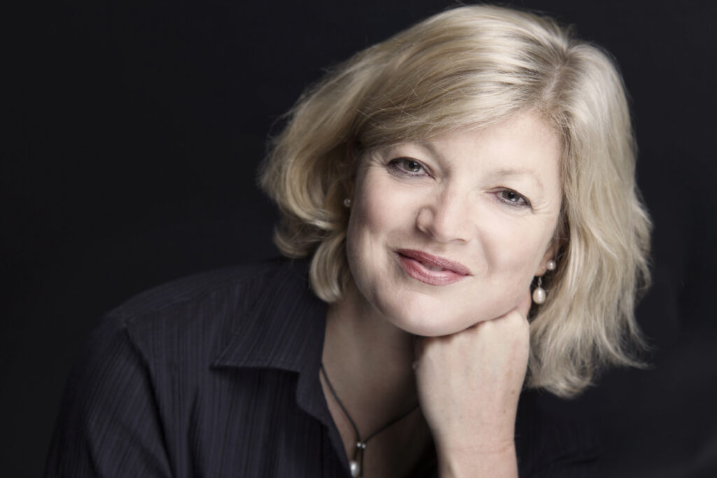 A smiling, fair-haired woman faces the camera, leaning on her hand to the right of the frame, a dark background behind her.