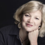 A smiling, fair-haired woman faces the camera, leaning on her hand to the right of the frame, a dark background behind her.