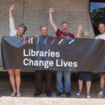 Five smiling people hold a black banner with white writing saying Libraries Change Lives. Rainbow coloured lines surround the words.