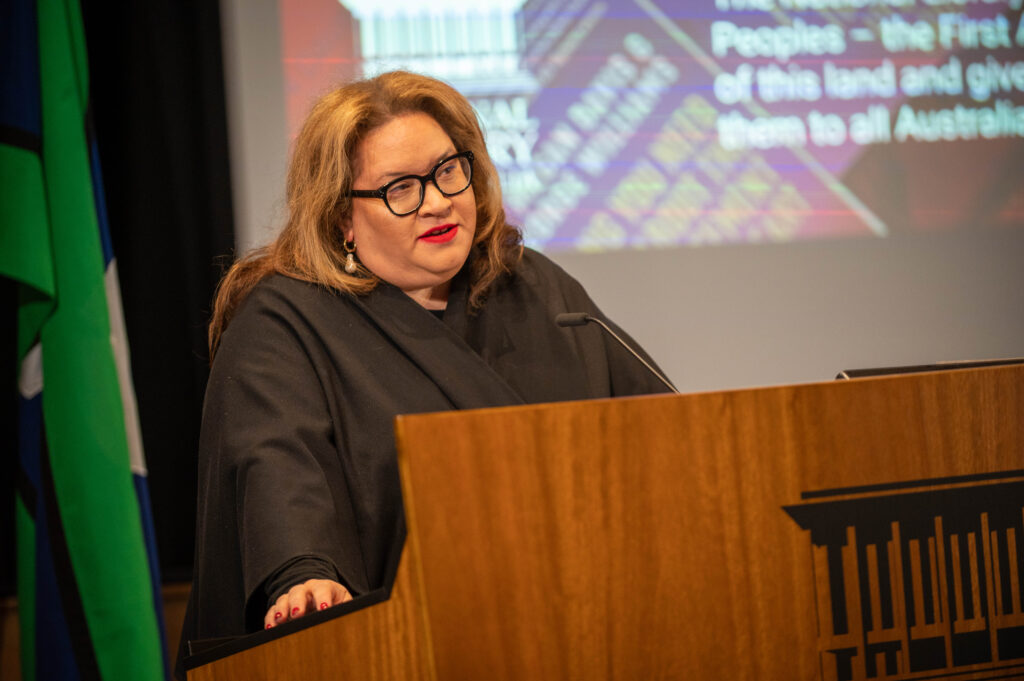 A woman with long hair, wearing black rimmed glasses , red lipstick and drop pearl earrings and a black academic gown, lectures at a podium. A Torres Strait Islands flag and slide mentioning 'Peoples - the first of this land' are visible in the background.