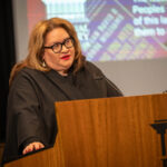 A woman with long hair, wearing black rimmed glasses , red lipstick and drop pearl earrings and a black academic gown, lectures at a podium. A Torres Strait Islands flag and slide mentioning 'Peoples - the first of this land' are visible in the background.