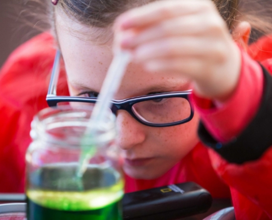 A young girl wearing glasses and a red top extracts green liquid from a glass jar using a dropper