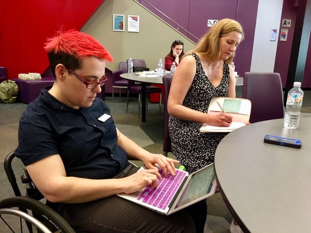 A person with bright red hair taps on their laptop computer from their wheelchair. Next to them, a blond women in a black and white dress write long-hand in a note book