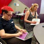 A person with bright red hair taps on their laptop computer from their wheelchair. Next to them, a blond women in a black and white dress write long-hand in a note book