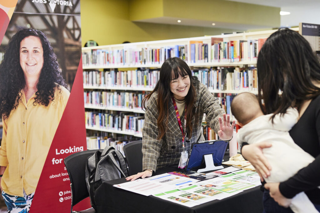 A woman smiles across a desk at a mother and babe in arms against a backdrop of library bookshelves