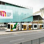Image of the facade of the Melbourne Convention and Exhibition Centre with a white, green and yellow tram passing in front of it.