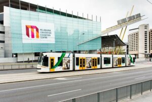 Image of the facade of the Melbourne Convention and Exhibition Centre with a white, green and yellow tram passing in front of it.