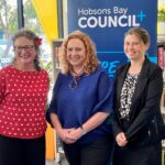 Three women stdn in front of a Hobsons Bay Council Banner. Angela is wearing red with a flower in her hair, Minister Horne is wearing royal blue and an artistic necklace, Diana Grima is wearing black and white