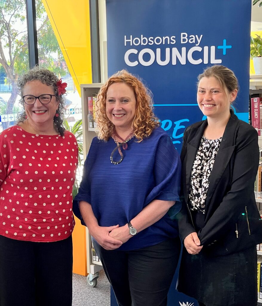 Three women stdn in front of a Hobsons Bay Council Banner. Angela is wearing red with a flower in her hair, Minister Horne is wearing royal blue and an artistic necklace, Diana Grima is wearing black and white