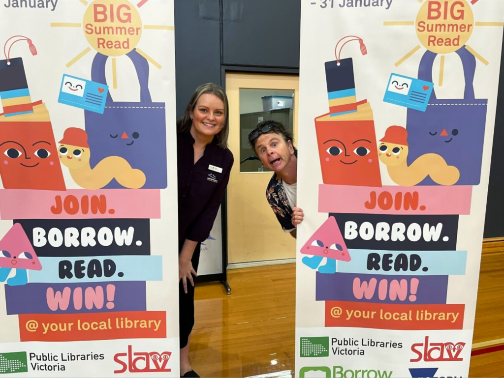 a woman and a man peek out from behind banners with graphic images of a sun saying Big Summer Read and a stack of books with Borow, Read, Win on the spines