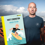 On the left is a copy of the picture book Surfing in the Dark showing an illustration of a man surfing on a yellow board on a blue wave. Behind the book is real-life blind surfer Matt Formston wearing a blue t-shirt, standing against a cloudy sky.
