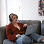 A woman reclines on a coach in her house, looking at a laptop and a pair of big headphones on.