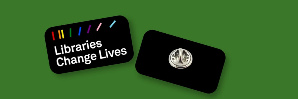 Two rectangular black pins on a green background. The left pin features multicoloured vertical lines along the top and the white text “Libraries Change Lives.” The right pin shows the back side with a silver butterfly clutch fastening.